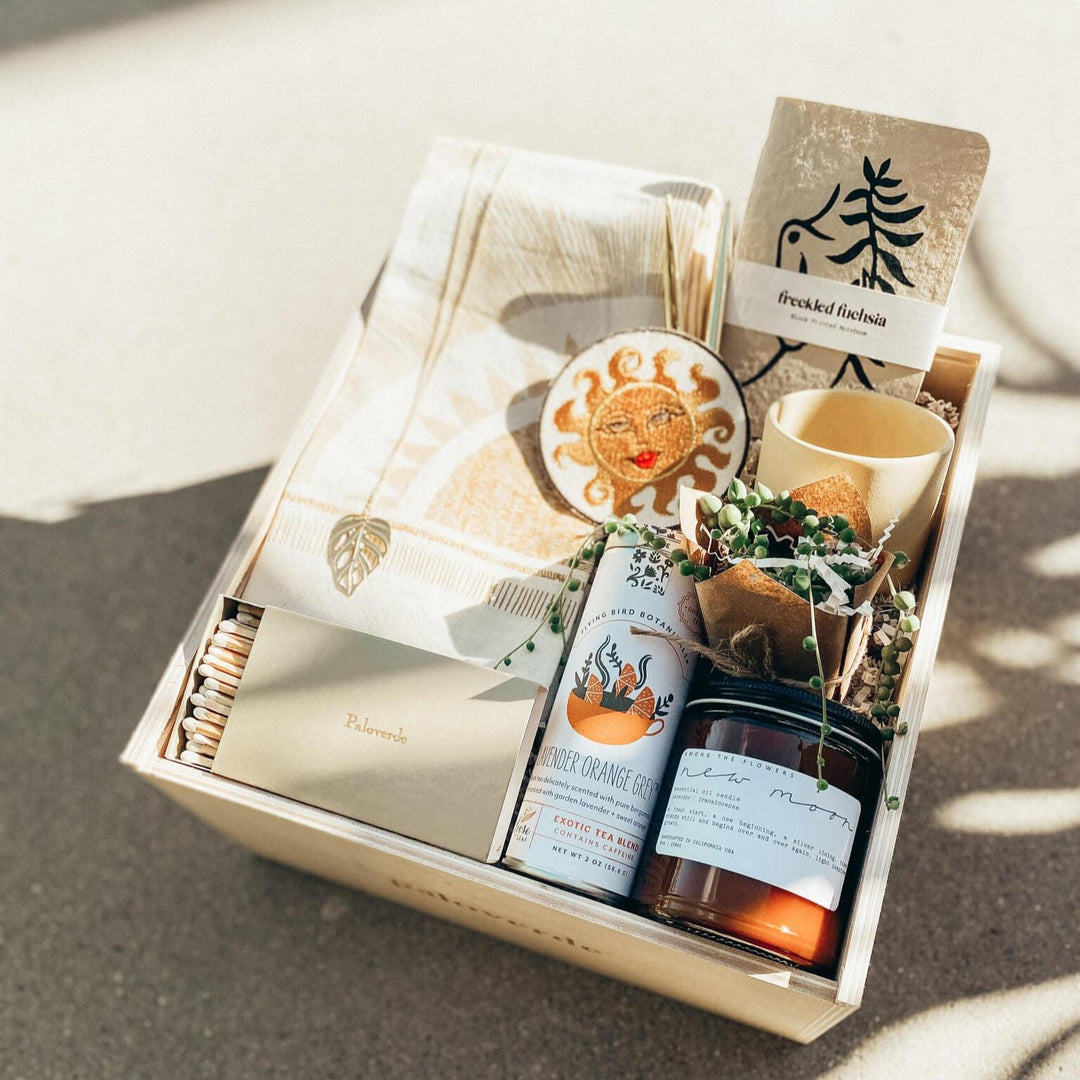 A custom Paloverde gift box sits in the dappled shadow of a plant and filled to the brim with gifts including tea tin, candle, gold matchbox, monstera necklace, sun patch, string of pearls plant and more.