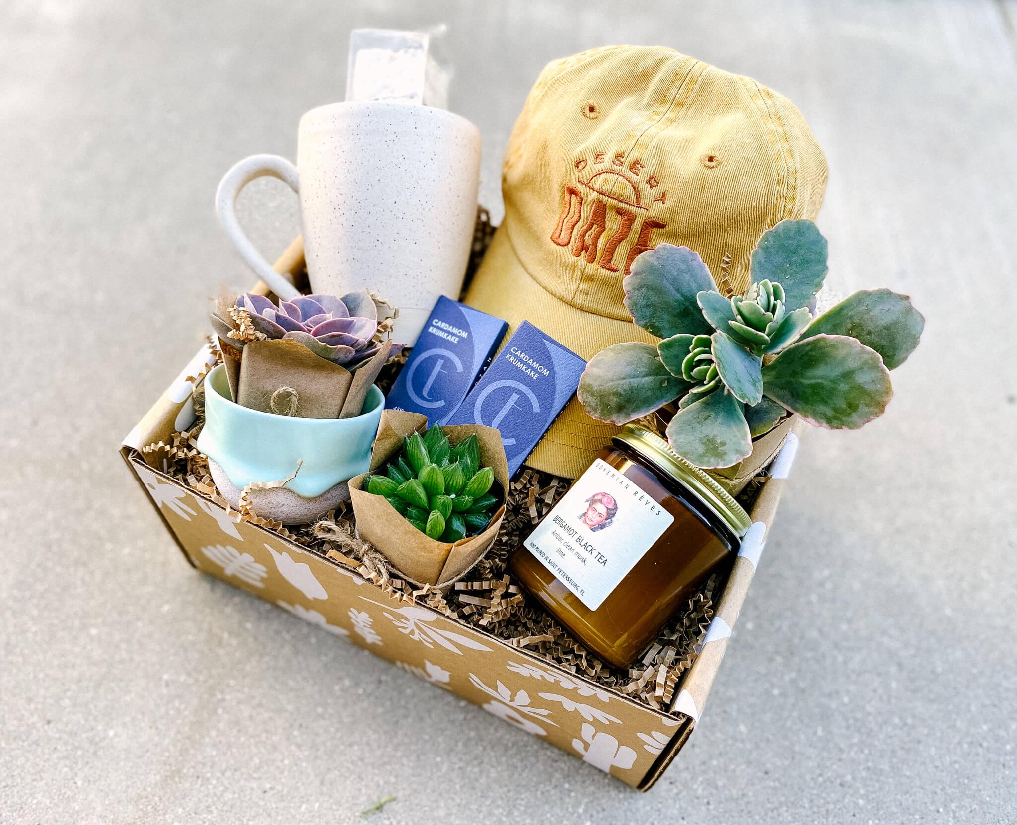 Custom built kraft gift box mailer filled with three succulent plants, a candle, cardamom kumkake chocolate bars, spreckled white mug, mint cappuccino cup, and a yellow hat with Desert Daze embroidery